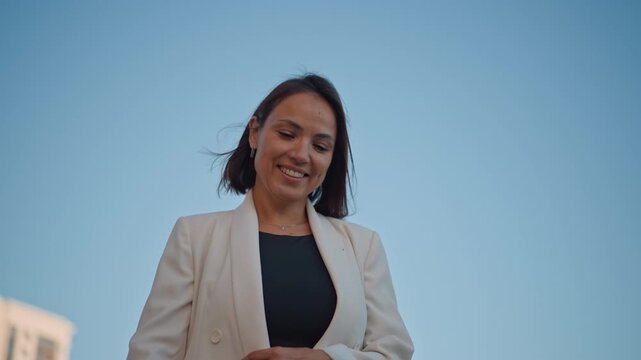 Young professional woman walking outdoors in modern city light, smiling with windblown hair, relaxed shoulders, and lively forward motion that suggests confidence and approachable energy