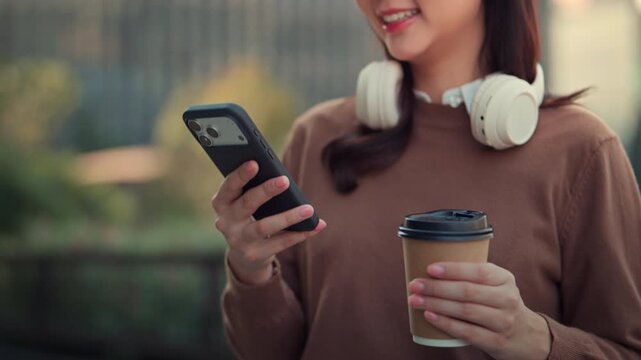 Happy young Asian woman using smartphone while holding takeaway coffee with headphones around neck, modern urban lifestyle and mobile connectivity