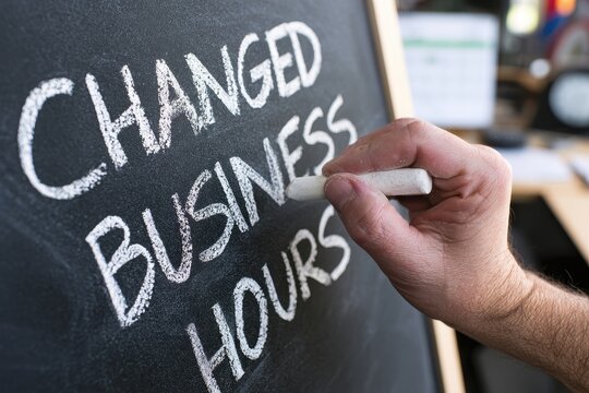 Hand writes CHANGED BUSINESS HOURS on a chalkboard in a bright office