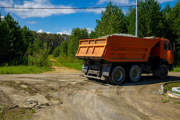 Obraz premium Orange dumper truck loaded with gravel standing on dirt road side in sunny weather. Concept: aggregates supply industry