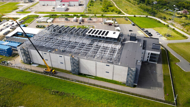 Aerial view showing a new data center building under construction with a large crane. Steel roof framework. Cooling systems. And an electrical substation in the background
