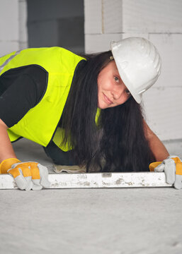 Construction worker at new house building site, kneeling, spirit level on ground in front of her, looking into camera, bare wall behind