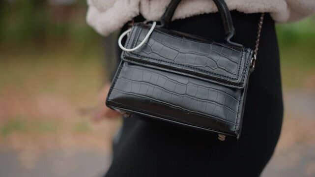 Side-waist close view of small black croc-textured leather handbag swinging at hip while person walks, with gloved hand near handle and chain strap as metal details catch soft overcast daylight
