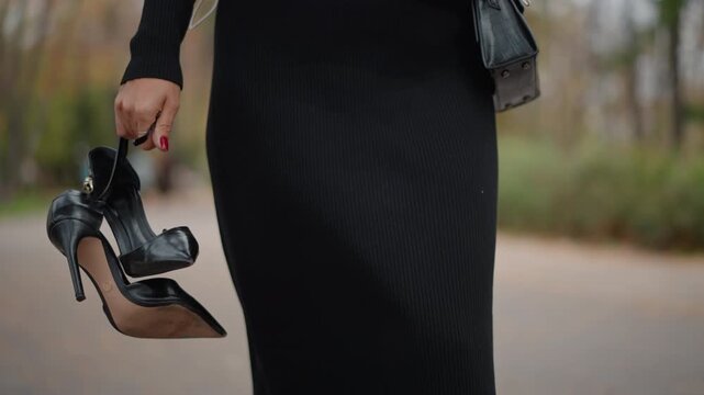 Lower-body view of woman walking along outdoor tree-lined path while holding black high-heeled shoes by straps at her side; shoes hang downward and swing slightly, indicating motion and transition