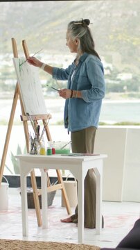 Vertical video: Stepping to easel, woman in denim selecting brushes, painting green marks at window