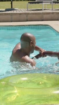 Vertical video: Reaching shirtless adult supporting shirtless teen playing in home pool, green tube