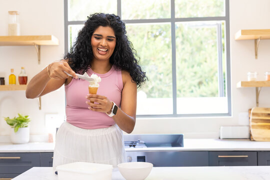 Indian woman in pink top scooping ice cream into cone with scoop at kitchen counter smiling