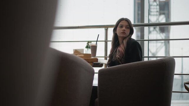 Young woman seated behind cushioned chairs in modern caf lounge turns head upward and left beside bright window, showing attentive reaction near wooden table with drink, soft daylight, and calm