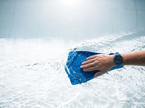A swimmer's hand wearing a blue sports watch propels through clear swimming pool water using a blue training paddle, creating dynamic splashes and ripples in bright sunlight