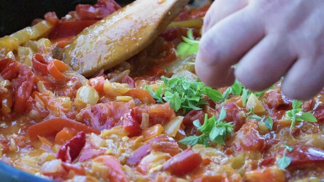 Hand sprinkling fresh green herb leaves over a bubbling tomato and pepper stew in a frying pan. Finishing touches to a healthy homemade meal.