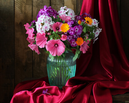Colorful lavatera and phlox bouquet in glass vase with red satin fabric and rustic wooden background