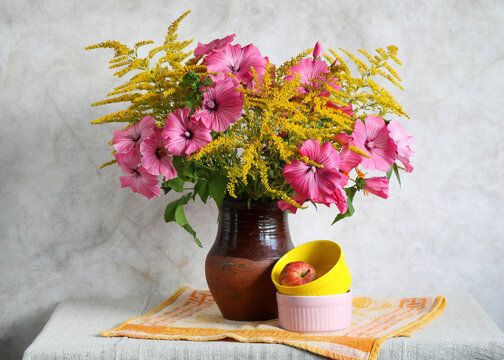 Bouquet of pink lavatera and goldenrod flowers in ceramic vase with apple and colorful bowls on table