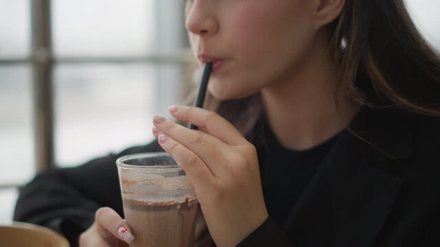 Young woman brings iced coffee to lips while adjusting straw with hand, relaxed expression, slight mouth opening, and soft indoor window light creating calm casual lifestyle mood, with woman, coffee
