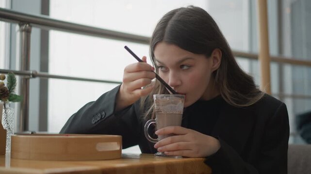 Young woman seated at wooden table in modern cafe lifts straw to mouth and stirs iced coffee with spoon, showing relaxed thoughtful expression, soft daylight, candid upper body view, cozy urban