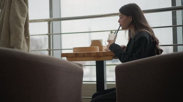 Young woman seated at round wooden cafe table brings cup with straw to her lips while gazing out large window, mid-sip motion shown by bent elbow and raised cup; calm, introspective mood conveyed by
