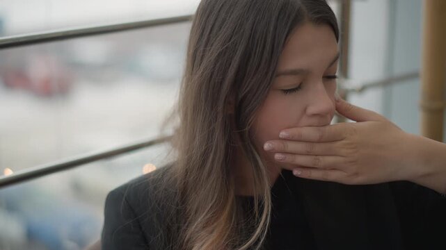 Close view of young woman seated beside window with urban railing, turning head aside and touching mouth while gazing downward, showing quiet contemplation, subtle emotion, soft daylight, and candid