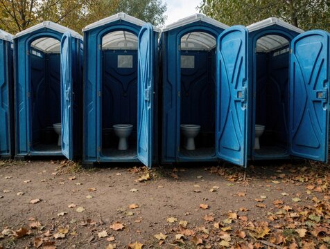 Blue portable toilets stand in a row with doors open. Outdoor temporary facilities on dusty ground with fallen leaves. Several vacant units ready for use.