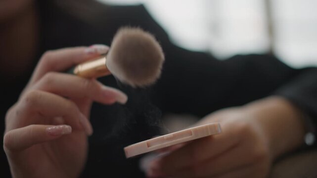 Tight side-view close-up of hands holding fluffy makeup brush above pink compact with visible sweeping motion and powder clinging to bristles, creating soft touch-up action with calm indoor light