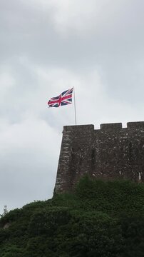 Union Jack flag rippling above weathered stone battlement, ivy on hillside, under muted overcast sky, with historic castle wall, fortress detail, textured masonry, and wind-driven movement