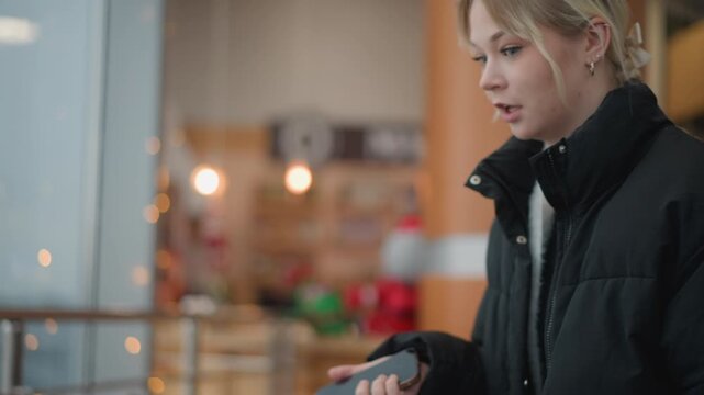 Young woman in dark coat sits on lounge chair inside cozy caf or shop interior, sampling small item between fingers while looking upward with thoughtful expression, warm light, soft blur and
