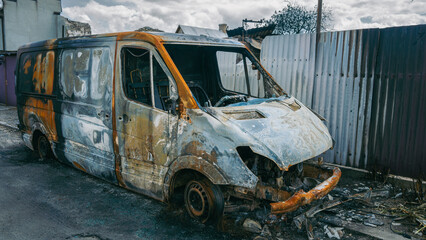 Obraz premium Side view of a completely burnt and destroyed delivery van in a war zone after military conflict, damaged fence background, war consequences concept