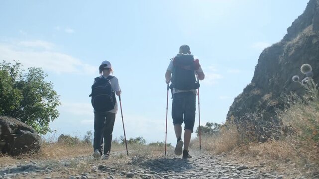 Low angle camera follows teenage brother and sister hiking along a dirt road with backpacks and trekking poles. Wearing caps for sun protection, outdoor hobby and leisure