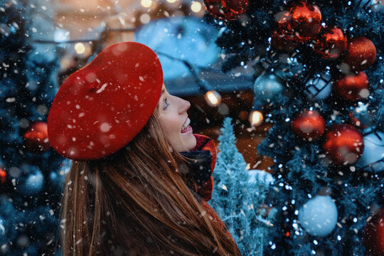 Woman in red hat smiles while looking up at holiday decorations during snowfall