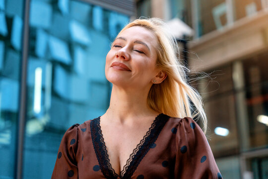 Woman stands outside a modern building with light reflections on glass and smiles while enjoying fresh air during a sunny day