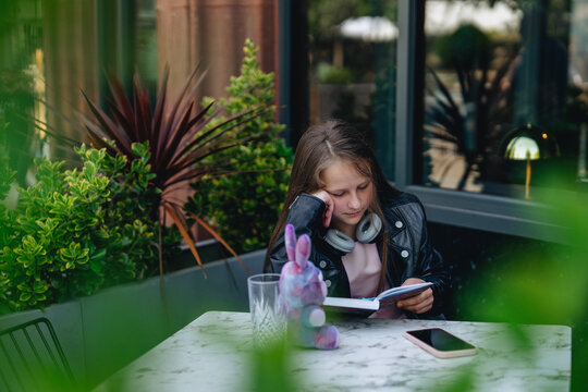 Girl reads book at cafe with plants and soft light in background during afternoon