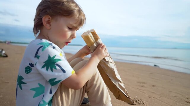 A happy toddler boy sits on warm sand, savoring a fresh baguette with pure joy. This little one enjoys a delicious snack outdoors, capturing a moment of peaceful contentment by the seaside.