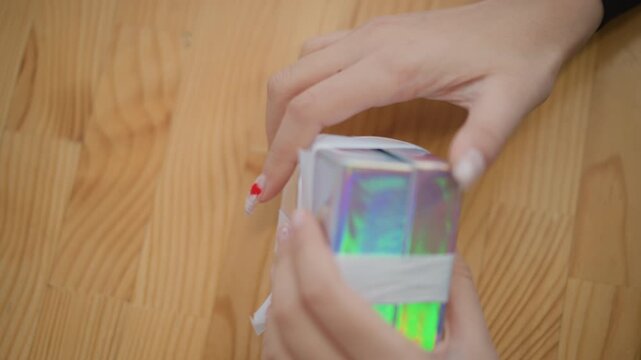 Close top view shows adult hands completing gift wrapping on small square box, pressing shiny lid, smoothing ribbon, folding edges, and aligning corners over rustic wooden table with soft shadow and