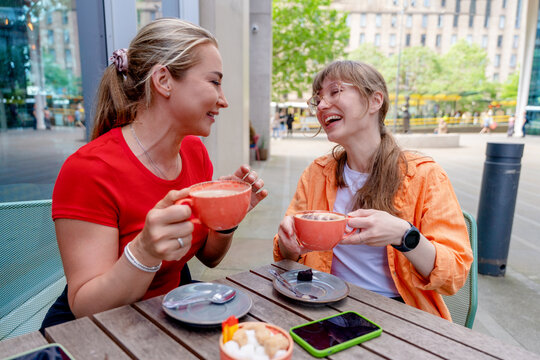 Two friends enjoy hot drinks at an outdoor cafe in a city during the day while laughing and talking together
