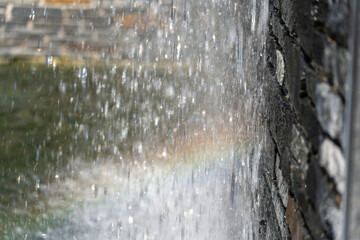 Obraz premium Water cascades down a stone wall in a fine mist that catches a soft rainbow. Droplets sparkle against the dark textured stones in this close-up.