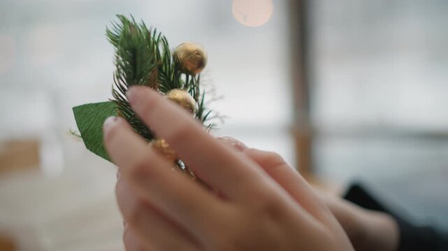 Close-up shows hands adjusting, placing, and securing holiday centerpiece with pine sprig, cedar branch, textured pinecone, gold berries, and green foliage, creating warm festive display