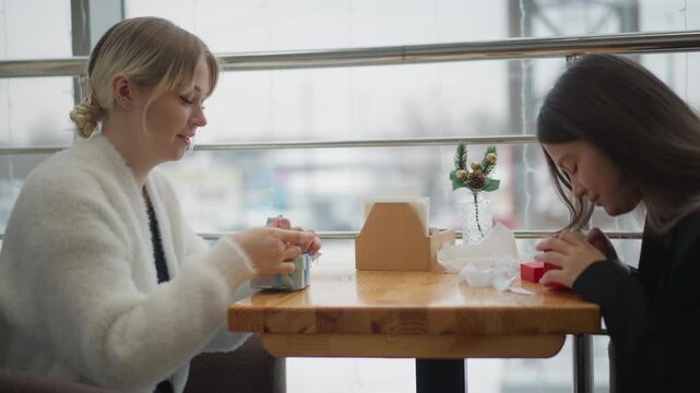 Two women sit opposite each other at wooden cafe table, exchanging small items while talking, smiling, and sipping drinks in soft daylight beside window, creating relaxed social atmosphere