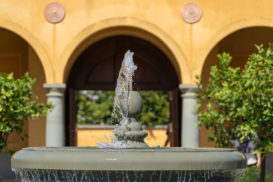 A tall fountain jet sprays water high above a stone basin framed by yellow Italianate archways. Potted citrus trees flank the courtyard on either side.