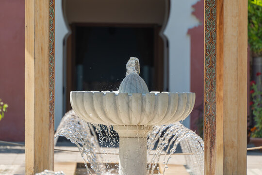 A shell-shaped stone fountain sprays water beneath a wooden pergola framing an ornate archway. Decorative tilework columns flank the bubbling water feature.