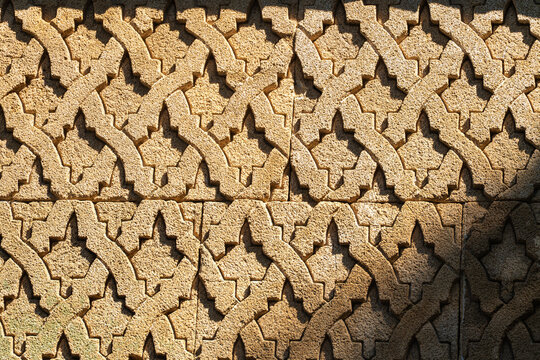 Intricate interlocking shapes are carved into a golden sandstone wall panel. Warm sunlight casts deep shadows across the repeating geometric pattern.