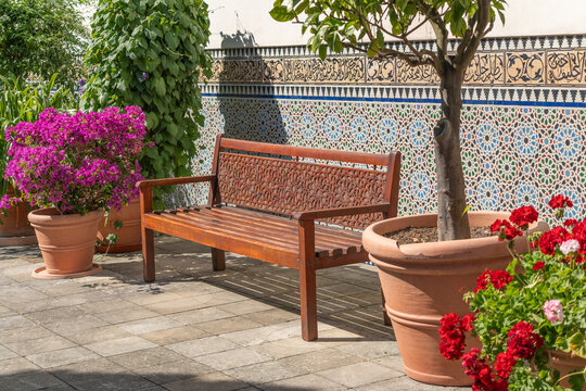 A carved wooden bench sits on a stone patio surrounded by vivid magenta bougainvillea and red geraniums in terracotta pots. An intricate mosaic tile wall rises behind the peaceful seating area.