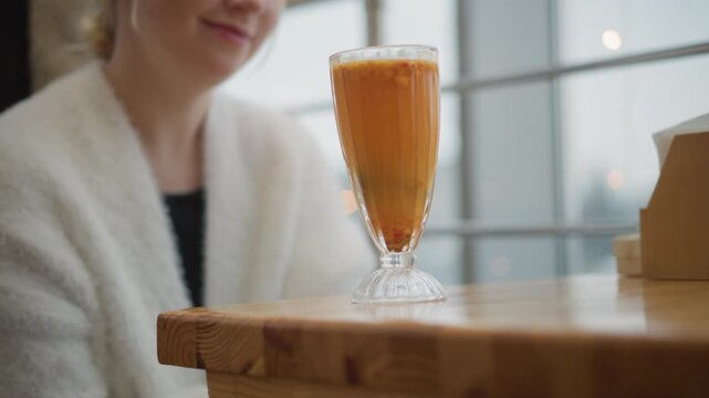 Ornate goblet of orange beverage rests on rustic wooden cafe table while blurred adult woman sits behind, creating warm indoor scene with condensation, gentle ripple, soft light, relaxed mood
