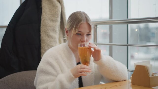 Young woman seated at wooden cafe table sips warm beverage from glass with both hands, lips touching rim while head tilts slightly forward and soft daylight from window shapes calm urban lifestyle