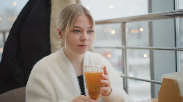 Young woman in wool sweater lifts glass of orange beverage to lips, taking calm sip beside bright caf window, with relaxed expression, soft daylight, cozy winter mood, gentle hand movement