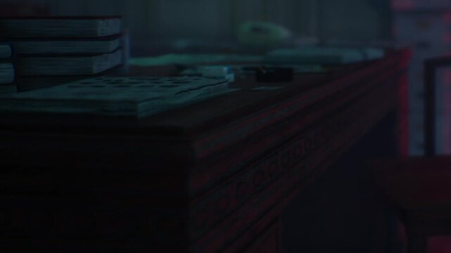 A wooden desk is shown with stacks of books and papers scattered on the surface. The light is low, creating shadows in the room. An old phone is placed on the desk.