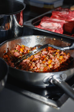 Browning minced meat with diced carrot and celery in stainless steel pan, spatula stirring soffritto on stove with raw beef tray blurred in background