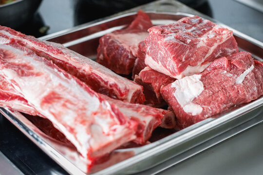 Raw bone-in beef short ribs and diced beef chunks in stainless steel tray on kitchen counter, close-up view of butchered meat