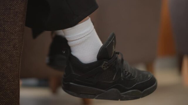 Low-angle close view shows seated feet in black sneakers and white socks gently swinging, with relaxed posture, casual indoor setting, soft light, and subtle motion creating quiet everyday comfort