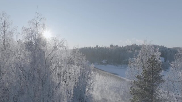Aerial drone footage flying over frozen river with sun glare in winter forest landscape