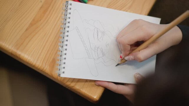 Right hand draws geometric shapes and guide lines with pencil on spiral sketchbook resting on wooden desk, fresh strokes and angled wrist showing active creative work in soft light