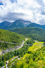 Obraz premium Highline 179 connects castle ruins with a Roman fortress. Visitors walk across the bridge over a wide river gorge. The scene shows mountain views and green landscape under a cloudy sky.