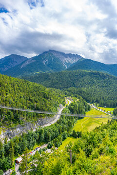 Highline 179 connects castle ruins with a Roman fortress. Visitors walk across the bridge over a wide river gorge. The scene shows mountain views and green landscape under a cloudy sky.